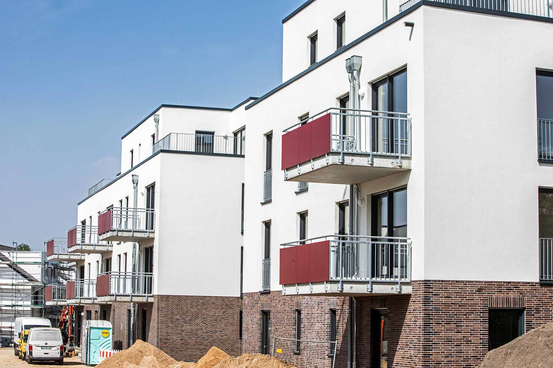 Neubau mit weißer Putzfassade, Klinkersockel und roten Balkonbrüstungen vor blauem Himmel