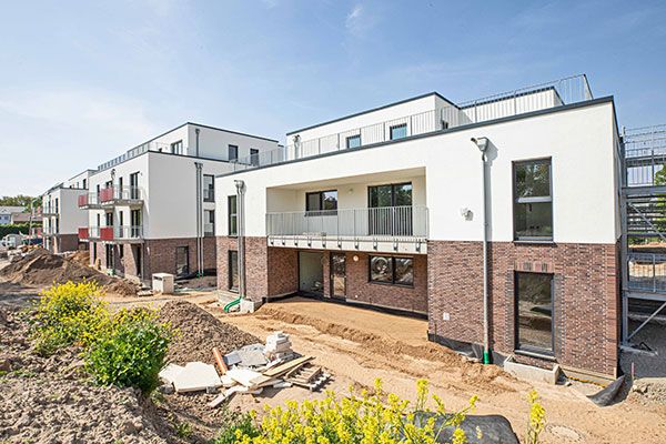 Neubau mit weiß verputzter Fassade, Klinkersockel und Balkon, im Vordergrund gelb blühende Sträucher