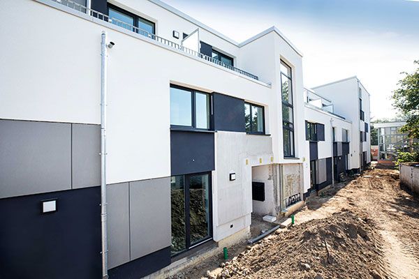 Neubau mit weißer Putzfassade und dunklen sowie grauen Fassadenplatten im Erdgeschoss, davor ein Sandweg