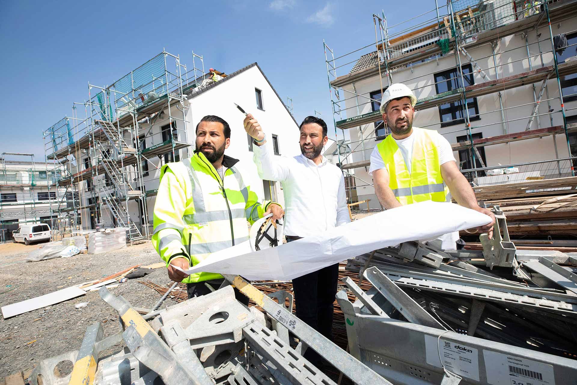 Drei Männer in Warnwesten besprechen einen Bauplan vor einem Gerüst auf einer Baustelle