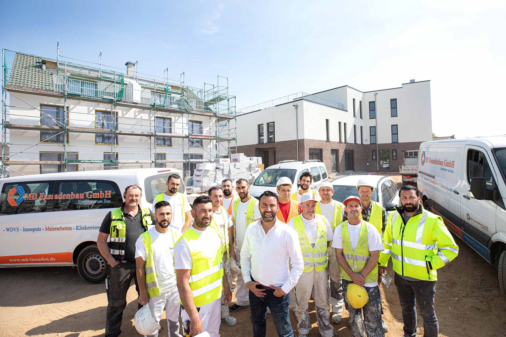 Gruppenfoto mehrerer Mitarbeiter in Warnwesten auf einer Baustelle vor einem Firmenfahrzeug der MD Fassadenbau GmbH